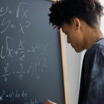 Side view of African American learner making math calculations on blackboard during lesson in classroom